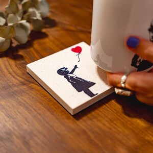 May include: A square off-white coaster featuring a black silhouette graphic of a person reaching toward a red heart-shaped balloon. The coaster is positioned next to a white ceramic mug that displays a matching black silhouette design.
