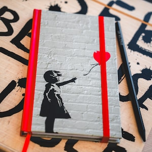 Könnte beinhalten: Ein Notizbuch mit rotem Band und einem grauen Ziegelmauer-Design. Der Umschlag zeigt eine schwarze Silhouette eines Mädchens, das nach einem roten herzförmigen Ballon greift. Der Text "Believe" ist in Schwarz auf der Holzoberfläche geschrieben.