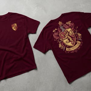 May include: Two maroon t-shirts. One shirt features a small gold and red crest on the chest. The other shirt displays a larger crest on the back, with a lion, helmet, and the word "Gryffindor".