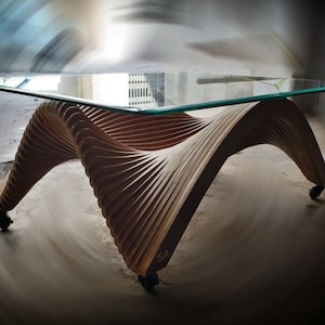 May include: A modern coffee table with a glass top and a unique, wavy wooden base. The base is made of multiple layers of wood that are curved and layered to create a flowing, organic design. The table is on wheels and has a signature "SA" carved into the wood.