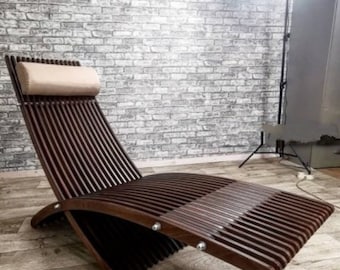 Op de afbeelding: Een donkerbruine houten chaise longue met een gebogen rugleuning en een gepolsterde hoofdsteun. De chaise longue is gemaakt van houten latten en heeft een modern ontwerp.