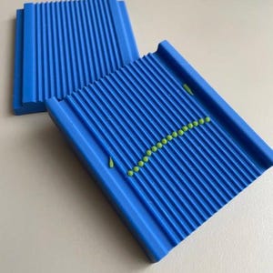 May include: Two rectangular blue plastic gnocchi boards featuring parallel ridges across the surface. One board displays a curved line of small, round, light green dough pieces arranged along the grooves, with two additional pieces placed separately.