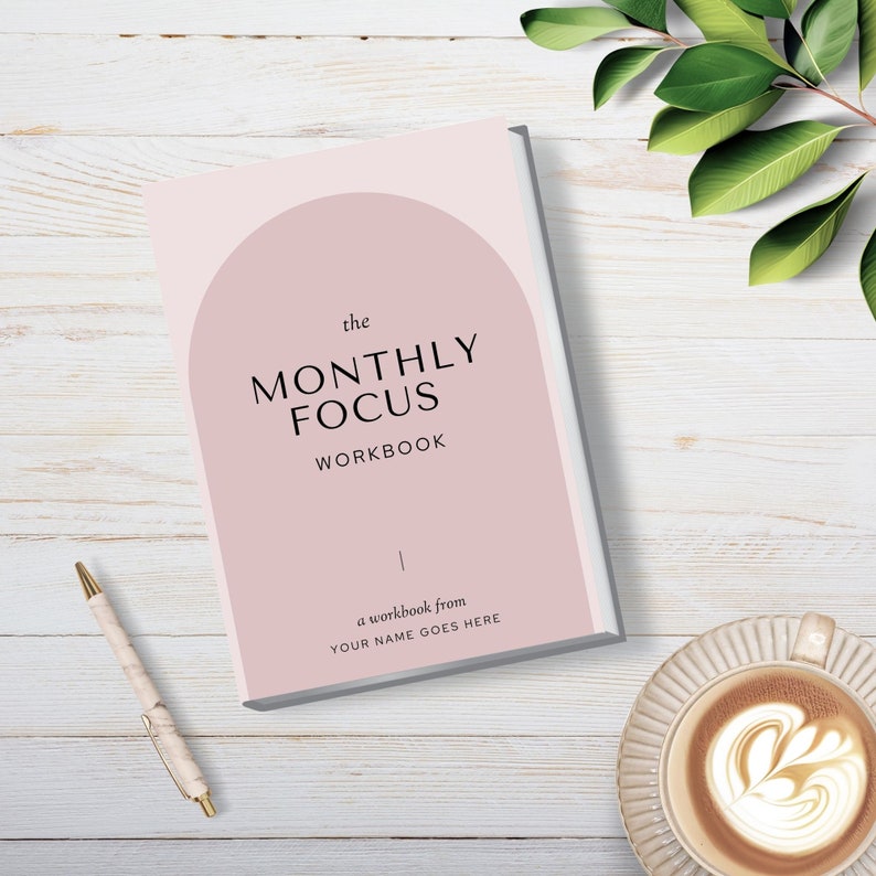 May include: A pink and white workbook with the text "the MONTHLY FOCUS WORKBOOK" and "a workbook from YOUR NAME GOES HERE" on a white wooden surface.