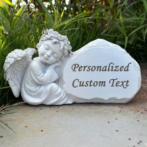 Könnte beinhalten: Weiße Keramikengelfigur mit Flügeln, die mit dem Kopf auf einem weißen Stein schläft, auf dem der Text "Personalized Custom Text" eingraviert ist.