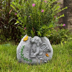 Könnte beinhalten: Ein grauer Steintopf in Form eines schlafenden Hundes mit Flügeln. Der Topf hat einen kleinen Schmetterling an der Seite und ist mit rosa Blumen gefüllt.