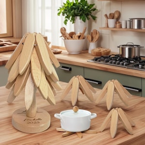 May include: A collection of wooden kitchen trivets and a decorative tree-shaped object. The trivets are made of light-colored wood and have a petal-like design. A white pot with a lid sits on one of the trivets. The tree-shaped object has a round base with the name "Michelle 2025" engraved.
