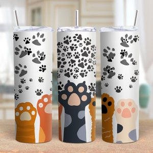 May include: Three stainless steel tumblers with paw print designs. The tumblers are white with black, brown, and gray paw prints. The tumblers have lids and straws.