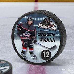 Puede incluir: Disco de hockey negro con una imagen circular de un jugador de hockey con uniforme negro y gris. El disco tiene el texto "Phantoms" y "2025-2026 U14AAA" y el número "12". Otro disco es visible en el fondo.