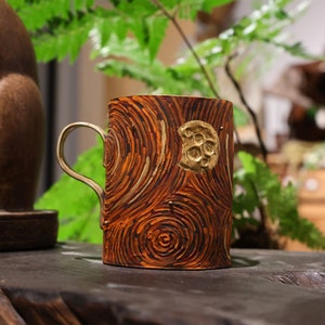 May include: A brown ceramic mug with a gold design of swirling lines and a crescent moon. The mug has a handle and is sitting on a wooden surface.
