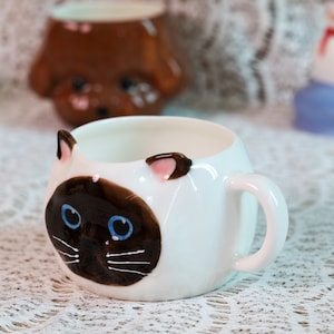 Op de afbeelding: Witte keramische mok met een bruin en wit kattengezichtontwerp. De kat heeft blauwe ogen en roze oren. De mok heeft een handvat.