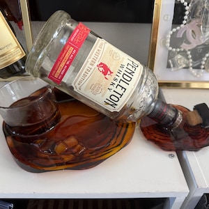 Peut inclure: Une bouteille de whisky Pendleton en verre transparent avec une étiquette rouge et blanche posée sur un plateau en résine ambrée foncée de forme organique. Le plateau présente une empreinte circulaire pour un verre et un support moulé.