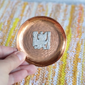 Könnte beinhalten: Eine kleine, gehämmerten Kupferschale mit einer silberfarbenen, stilisierten Figur in der Mitte. Die Schale hat eine leicht konkave Form und eine polierte, reflektierende Oberfläche. Der Hintergrund ist ein gewebter Textil mit gelben und weißen Fäden.