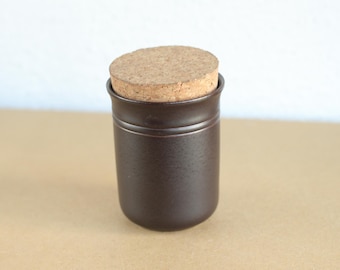 May include: A small, dark brown ceramic jar with a cork stopper. The jar has a cylindrical shape with a slightly rounded base and a horizontal groove near the top. The cork is light brown and fits snugly into the jar's opening.