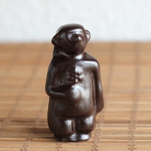 May include: A small, dark brown ceramic figurine of a person with their hands clasped in front of them. The figurine is standing on a woven bamboo mat.