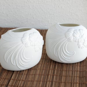 May include: Two white ceramic vases with a floral design. The vases have a rounded shape with a wide opening at the top. The floral design is embossed on the front of the vases. The vases are sitting on a brown woven mat.