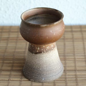 Könnte beinhalten: Eine braune und weiße Keramikvase mit einer breiten, abgerundeten Oberseite und einem schmalen Boden. Die Vase hat eine gesprenkelte Glasur und ein rustikales Aussehen.
