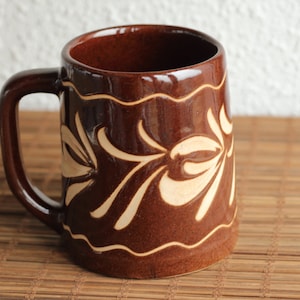 May include: A brown ceramic mug with a white floral design. The mug has a handle and a rounded shape.