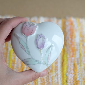 Könnte beinhalten: Weiße herzförmige Schmuckschatulle mit Blumendesign mit rosa und lila Tulpen. Die Keramikbox hat eine glänzende Oberfläche. Die Tulpen haben grüne und hellblaue Blätter.