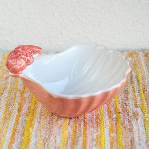 Op de afbeelding: Een keramische schelpvormige kom met een witte binnenkant en een koraalkleurige buitenkant. De kom heeft een gestructureerd, geribbeld ontwerp en een koraalkleurig kreeftenklauwdetail aan de zijkant. De kom staat op een geel en wit gestreept oppervlak.