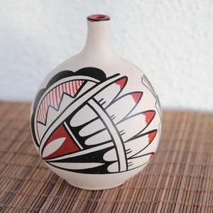 May include: Cream-colored ceramic vase with a rounded body and a narrow neck. The vase features a Native American-inspired design in black, red, and white. The design includes geometric shapes and feather-like elements. The rim is red.