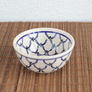 Könnte beinhalten: Eine kleine, runde Keramikschale mit weißem Grund und blauem Muster. Das Muster besteht aus miteinander verbundenen blauen Formen mit kleinen grünen Akzenten. Die Schale steht auf einer braunen, gewebten Matte.
