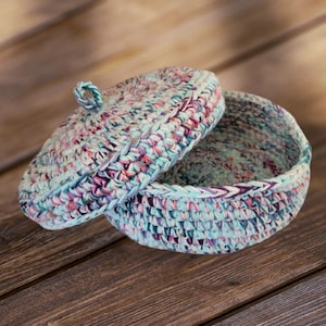 May include: A crocheted storage basket with a lid. The basket is made of a multi-colored yarn in shades of blue, pink, and white. The lid has a small loop handle.