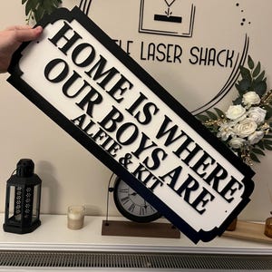 May include: Black and white wooden sign with the text "Home is where our boys are Alie & Kit".