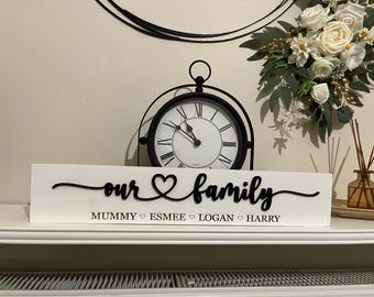 Può includere: Un cartello rettangolare in legno bianco con la scritta nera in rilievo "our family" e un'icona a forma di cuore al centro. Sotto la scritta, i nomi "MUMMY", "ESMEE", "LOGAN" e "HARRY" sono stampati in lettere maiuscole nere, separati da piccoli cuori.