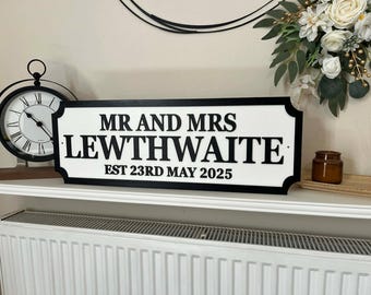 Può includere: Un cartello rettangolare bianco con bordo nero e scritte nere che recita MR AND MRS LEWTHWAITE EST 23RD MAY 2025. Il cartello presenta angoli decorativi intagliati ed è esposto su una mensola bianca.