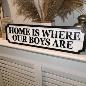 May include: White wooden sign with black trim and black lettering that reads "Home is where our boys are".