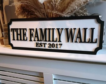 Può includere: Un cartello rettangolare con sfondo bianco e bordo nero. Mostra la scritta "THE FAMILY WALL EST 2017" in lettere nere in grassetto. È un elemento decorativo, forse per una casa.