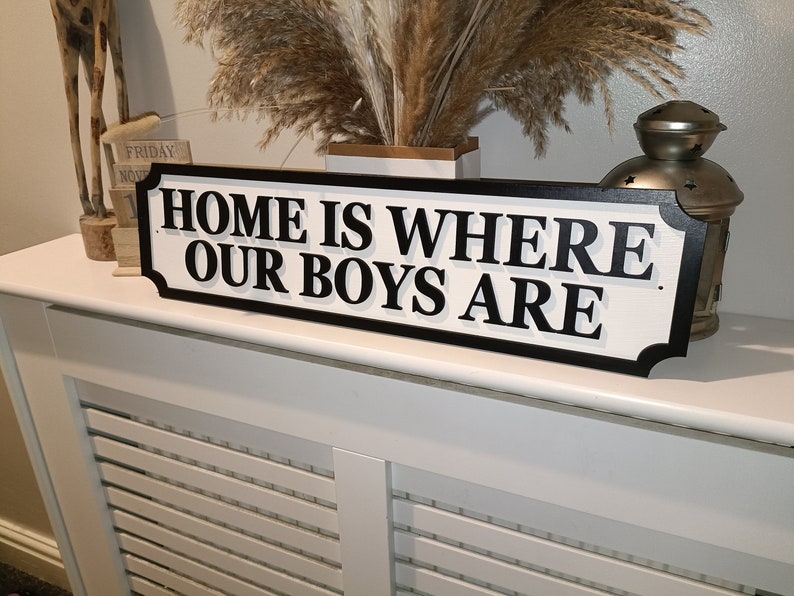 May include: White wooden sign with black lettering that reads "Home is where our boys are".