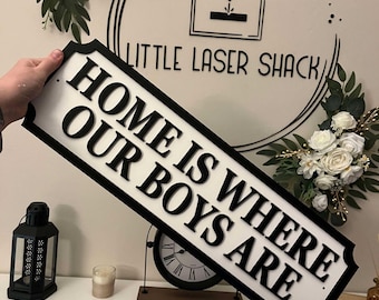 Può includere: Un cartello in legno bianco con lettere nere che recita "Home is where our boys are". Il cartello è decorato con un bordo nero e un bordo decorativo. Il cartello è tenuto da una mano.