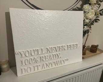 Può includere: Opera d'arte su tela bianca strutturata con la citazione "YOU'LL NEVER FEEL 100% READY. DO IT ANYWAY." L'opera è esposta su una superficie bianca, con una candela e fiori decorativi sullo sfondo.