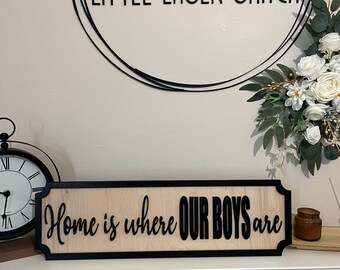 Può includere: Un cartello di legno con cornice nera e il testo "Home is where our boys are" in lettere nere.
