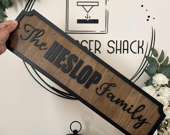 Può includere: Un'insegna rettangolare in legno con finitura a venature marrone scuro e bordo nero. L'insegna presenta il testo in rilievo nero "The HESLOP Family" in una combinazione di caratteri serif e script centrati sulla superficie.