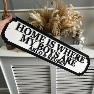 May include: White wooden sign with black lettering that reads "Home is where my boys are Arlo & Reuben".