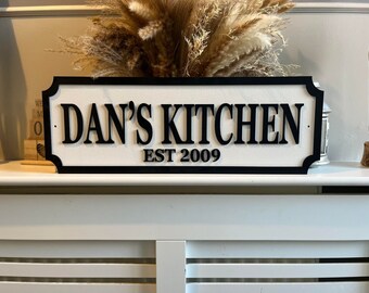 Può includere: Un cartello in legno bianco e nero con il testo "Dan's Kitchen Est 2009".