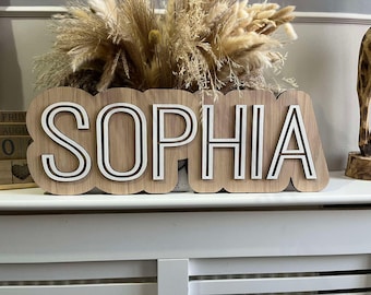 Può includere: Un cartello in legno con il nome "SOPHIA" in lettere bianche con contorno nero. Le lettere sono in rilievo e hanno un effetto 3D. Il cartello è in legno marrone chiaro e ha un aspetto rustico.