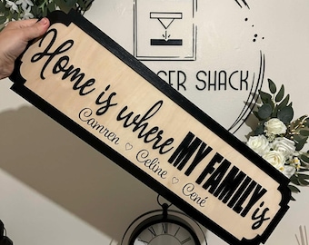 Può includere: Un cartello in legno con la frase "Home is where my family is" in lettere nere. Il cartello ha un centro in legno chiaro con un bordo nero. I nomi Camren, Celine e Cené sono sotto il testo principale. Il cartello è tenuto in mano.