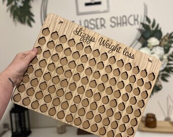Może przedstawiać: Drewniany tracker utraty wagi ze 100 kółkami do zaznaczania postępów. Tracker jest grawerowany tekstem "Lizzy's Weight Loss".