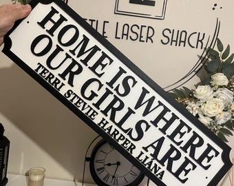 Può includere: Un cartello in legno bianco e nero con il testo "Home is where our girls are." I nomi Terri, Stevie, Harper e Liana sono elencati sotto il testo.