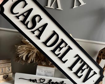 Può includere: Un cartello in legno bianco e nero con il testo "CASA DEL TEV" in un carattere decorativo. Il cartello è appeso a una parete con altre lettere decorative.