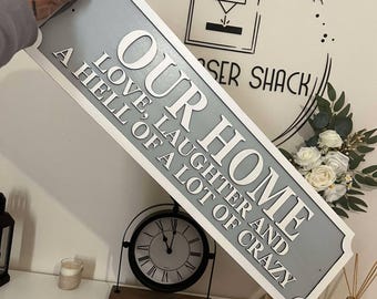 Può includere: Un'insegna rettangolare grigia con scritte bianche che recita "OUR HOME LOVE, LAUGHTER AND A HELL OF A LOT OF CRAZY". L'insegna è tenuta davanti a un muro con un orologio e decorazioni floreali.