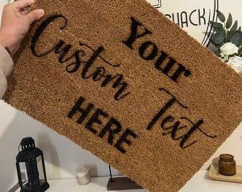 Può includere: Un tappetino per porta in fibra di cocco marrone con il testo "Your Custom Text Here" in lettere nere.