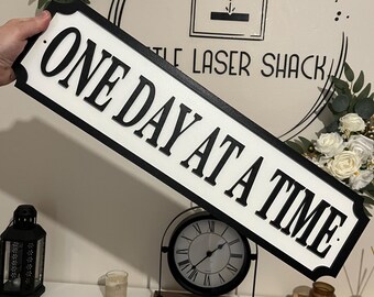Può includere: Un cartello in legno bianco e nero con il testo "ONE DAY AT A TIME" in un carattere decorativo. Il cartello è appeso a un muro con un orologio e una candela su uno scaffale sottostante.