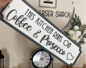 Può includere: Un cartello rettangolare in legno bianco e grigio con la scritta "THIS KITCHEN RUNS ON Coffee & Prosecco". Il cartello ha un bordo grigio e un simbolo a forma di cuore. Il cartello è una decorazione per la cucina.