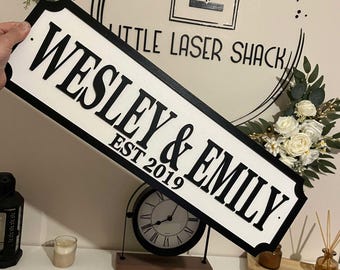 Può includere: Un cartello in legno bianco con lettere nere che recita "Wesley & Emily Est 2019". Il cartello è in stile segnale stradale con angoli arrotondati e un bordo nero.
