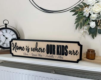 Può includere: Un cartello in legno con la frase "Home is where OUR KIDS are" in lettere nere. Il cartello include i nomi Olivia, Ronnie-Mae, Arabella e Albie. Il cartello è incorniciato in nero e poggia su una superficie bianca.