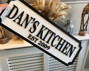 Può includere: Un cartello in legno bianco con lettere nere che recita "Dan's Kitchen Est 2009". Il cartello ha un bordo nero e un bordo decorativo.